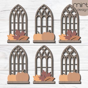 May include: Six wooden fall-themed decorations. Each features a gothic arch window design with pumpkins and fall leaves. The decorations are in shades of brown, orange, and tan. The brand name "mirta designs" is visible.