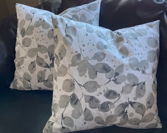 May include: Two throw pillows with a white background and a gray leaf pattern. The leaves have a watercolor effect and there are gray splatters on the fabric.