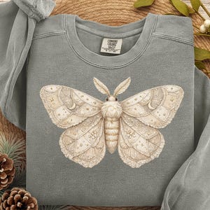 May include: Gray sweatshirt featuring a detailed illustration of a moth with intricate wing patterns. The moth is cream-colored with gold accents. The sweatshirt has long sleeves and a crew neck. The words "Your Brand Name" and "Gray" are visible.