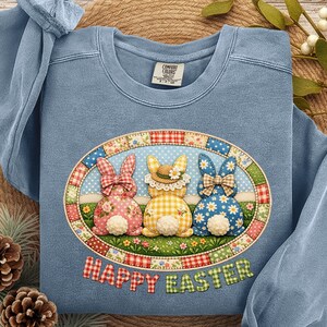 May include: A light blue sweatshirt with an oval design featuring three bunny rabbits with floral, gingham, and polka dot patterns. The words "HAPPY EASTER" are below the design. The sweatshirt is made of a soft material.