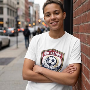 Può includere: Una maglietta bianca con un'emblema di pallone da calcio con la scritta "ONE NATION". Il design è a forma di scudo, con una corona d'alloro. La persona è appoggiata a un muro di mattoni.