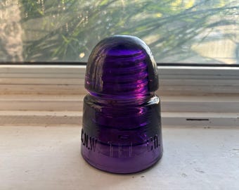 May include: A deep purple glass telegraph insulator with a rounded top and ribbed sides. The base features embossed lettering that reads C.N.W. TEL. CO. The object has a smooth, translucent finish and a wide, flat bottom for stable placement.