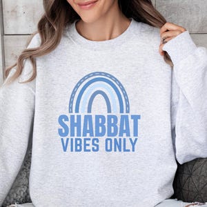 May include: A light gray crewneck sweatshirt featuring a blue graphic of a rainbow and the text SHABBAT VIBES ONLY printed in a bold sans-serif font. The garment has long sleeves, a ribbed neckline, and ribbed cuffs, providing a casual look.