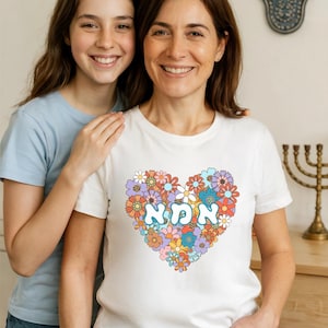 Könnte beinhalten: Zwei Personen, eine Mutter und ihre Tochter, tragen weiße T-Shirts mit einem farbenfrohen Blumenherz-Design. Das Herz zeigt das hebräische Wort "ima" in der Mitte, was "Mutter" bedeutet.