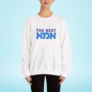 Könnte beinhalten: Weißes Sweatshirt mit dem Text "THE BEST NON" in blauem Glitzer-Schriftzug. Das Sweatshirt ist ein Rundhals-Stil und wird von einer Person getragen. Der Hintergrund ist hellblau.