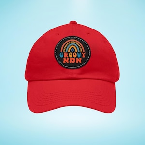 May include: A red baseball cap with a circular patch displaying a rainbow design and the words "GROOVY אמא" in a retro font. The patch includes a Star of David symbol. The cap is made of a durable fabric and has a curved peak.