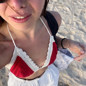 Op de afbeelding: Een handgemaakte gehaakte bikinitop in rood en wit. De top heeft een geschulpte rand en een halternek ontwerp. De afbeelding toont ook een witte broek en een strandomgeving.