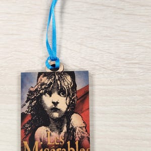 Puede incluir: Un marcapáginas de madera con una cinta azul atada. El marcapáginas presenta una ilustración en blanco y negro de una niña con el texto "Les Misérables" en letras doradas.