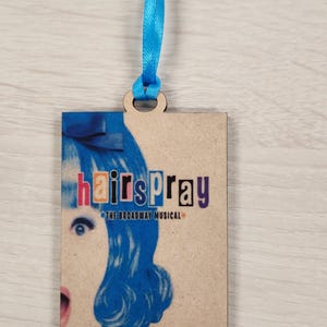 May include: A wooden ornament with a colorful image of a woman with blue hair and the text "hairspray the broadway musical".