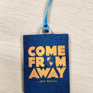 May include: A blue wooden ornament with a yellow and orange graphic that reads "COME FROM AWAY A NEW MUSICAL". The ornament has a blue ribbon attached to it.