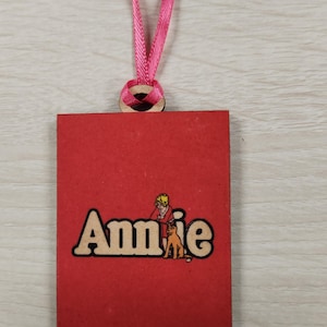 May include: A red rectangular wooden ornament with the name "Annie" in a cartoon font. The ornament features a cartoon image of a girl and a dog. A pink ribbon is attached for hanging.