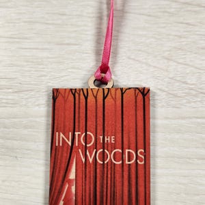 May include: A wooden bookmark with a red curtain and the text "INTO THE WOODS". A small figure of a girl in a red hood is visible behind the curtain.