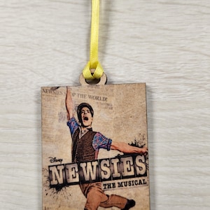 May include: A wooden ornament featuring a vintage-style poster for the Disney musical "Newsies." The poster depicts a young man in a brown and white plaid shirt, jumping in the air with his arms outstretched. The text "Disney Newsies The Musical" is printed in large, bold letters on the poster.