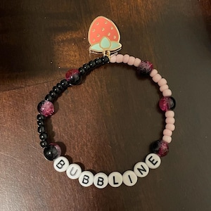 May include: A beaded bracelet with the word "BUBBLINE" spelled out in white letter beads. The bracelet features black and pink beads, with a strawberry charm at the top. The strawberry charm is red with green accents.