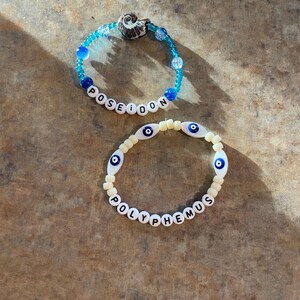 May include: Two beaded bracelets. One is turquoise with the word "POSEIDON" spelled out in white letter beads and a seashell charm. The other is cream-colored with blue eye beads and the word "POLYPHEMUS".