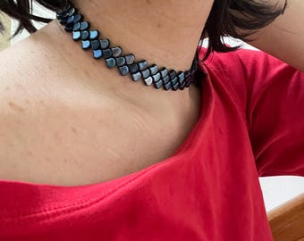 May include: A black choker necklace with a scale-like design. The necklace is made of small, iridescent, dark blue and gray pieces. The person in the image is wearing a red shirt.