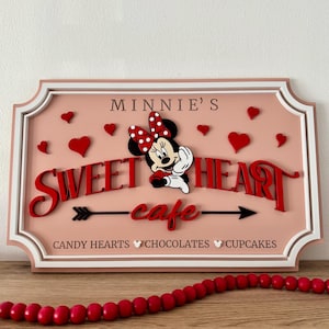 May include: A pink and white sign with a red and white Minnie Mouse illustration. The sign reads "Minnie's Sweetheart cafe" with a red arrow pointing to the right. The text "Candy Hearts Chocolates Cupcakes" is at the bottom of the sign.