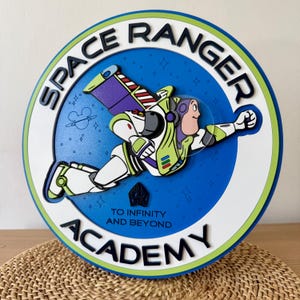 May include: A circular Space Ranger Academy sign with a cartoon astronaut in flight. The sign has a blue background with white and green borders. The text "SPACE RANGER ACADEMY" and "TO INFINITY AND BEYOND" are displayed.
