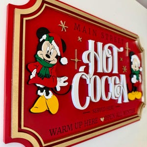 K&ouml;nnte beinhalten: Ein rotes Holzschild mit goldener Umrandung und einem wei&szlig;en 3D-Schriftzug "Hot Cocoa". Mickey Mouse und Minnie Mouse sind in Winterkleidung gekleidet und befinden sich auf beiden Seiten des Schildes. Das Schild lautet "* Main Street * Hot Cocoa * Served Here * Warm Up Here Open All Winter".
