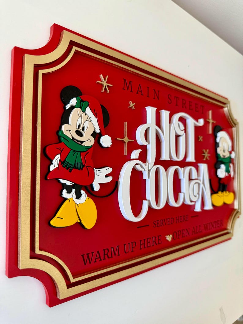 K&ouml;nnte beinhalten: Ein rotes Holzschild mit goldener Umrandung und einem wei&szlig;en 3D-Schriftzug "Hot Cocoa". Mickey Mouse und Minnie Mouse sind in Winterkleidung gekleidet und befinden sich auf beiden Seiten des Schildes. Das Schild lautet "* Main Street * Hot Cocoa * Served Here * Warm Up Here Open All Winter".