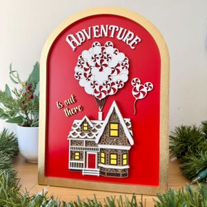 Könnte beinhalten: Ein mehrschichtiges Holz-Kunstwerk mit rotem Hintergrund und goldenem Rahmen. Das Design zeigt ein Haus mit einem Ballon-Cluster, einem Mickey-Mouse-förmigen Ballon und dem Text "Adventure is out there". Das Haus hat aufwendige Details.