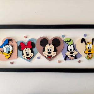 May include: A framed artwork featuring Disney characters Donald Duck, Minnie Mouse, Mickey Mouse, Goofy, and Pluto, each within a heart-shaped design. The characters are in vibrant colors, set against a white background, and framed in black.