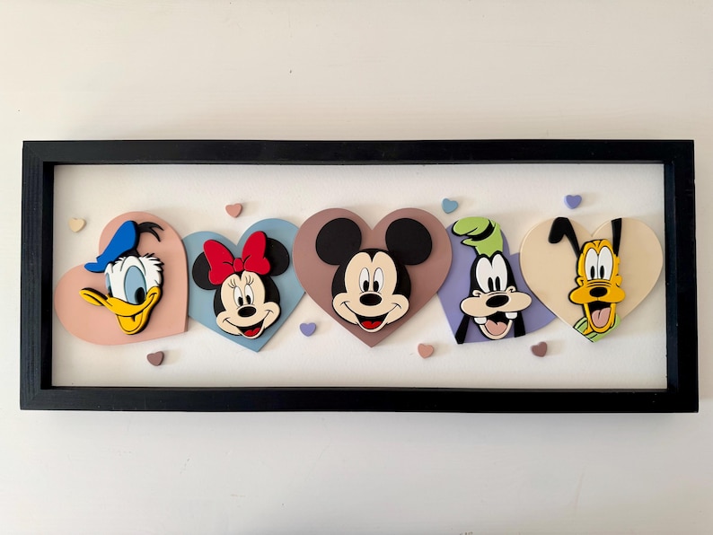 May include: A framed artwork featuring Disney characters Donald Duck, Minnie Mouse, Mickey Mouse, Goofy, and Pluto, each within a heart-shaped design. The characters are in vibrant colors, set against a white background, and framed in black.