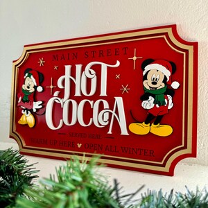 K&ouml;nnte beinhalten: Rotes Holzschild mit goldener Umrandung, auf dem Mickey Mouse und Minnie Mouse in Winterkleidung zu sehen sind. Das Schild lautet "Main Street Hot Cocoa Served Here Warm Up Here Open All Winter".
