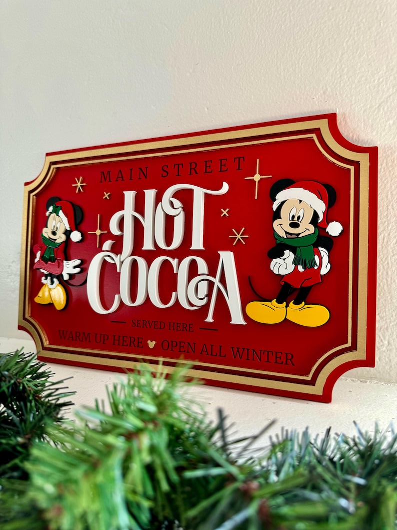 K&ouml;nnte beinhalten: Rotes Holzschild mit goldener Umrandung, auf dem Mickey Mouse und Minnie Mouse in Winterkleidung zu sehen sind. Das Schild lautet "Main Street Hot Cocoa Served Here Warm Up Here Open All Winter".