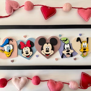 May include: A framed artwork featuring Disney characters Donald Duck, Minnie Mouse, Mickey Mouse, Goofy, and Pluto, each on a heart-shaped background. The frame is black, and the artwork is decorated with felt hearts and pom-poms.