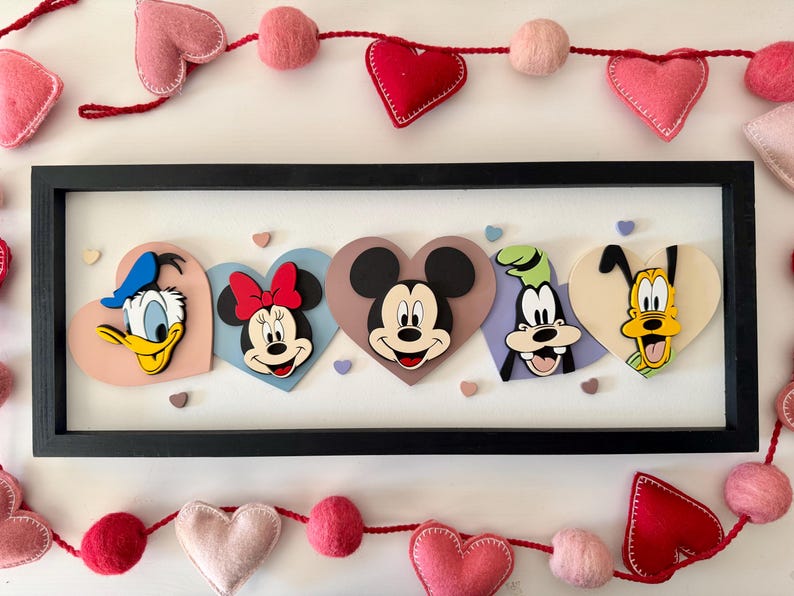 May include: A framed artwork featuring Disney characters Donald Duck, Minnie Mouse, Mickey Mouse, Goofy, and Pluto, each on a heart-shaped background. The frame is black, and the artwork is decorated with felt hearts and pom-poms.