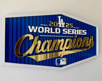 Könnte beinhalten: Ein gerahmtes Schild mit blauem Hintergrund und vertikalen Linien. Das Schild zeigt den Text "2025 World Series Champions" in Gold und Weiß, mit dem LA Dodgers-Logo. Der Rahmen ist weiß.