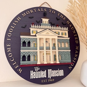 Könnte beinhalten: Ein rundes, violettes Wandschild aus Holz mit einem dreidimensionalen, geschichteten Modell eines weißen Herrenhauses mit verzierten blauen Geländern. Der Text "WELCOME FOOLISH MORTALS TO THE HAUNTED MANSION" steht am Rand, darunter "The Haunted Mansion EST 1969".