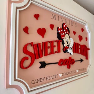 May include: A pink and white sign with a red and white Minnie Mouse character. The sign says "Minnie's Sweetheart Cafe" with an arrow pointing to the right. The sign also says "Candy Hearts, Chocolates, Cupcakes".