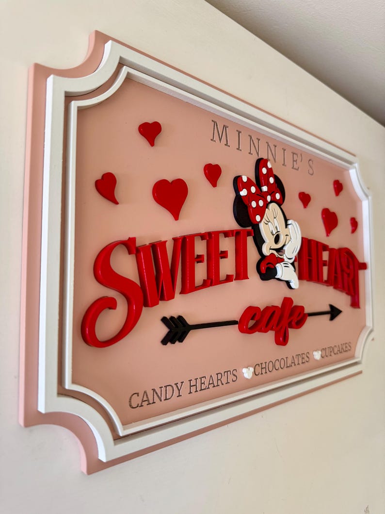 May include: A pink and white sign with a red and white Minnie Mouse character. The sign says "Minnie's Sweetheart Cafe" with an arrow pointing to the right. The sign also says "Candy Hearts, Chocolates, Cupcakes".
