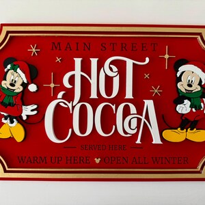 K&ouml;nnte beinhalten: Ein rotes Holzschild mit goldenem Rahmen, auf dem Mickey und Minnie Mouse in Weihnachtskleidung zu sehen sind. Das Schild lautet "Main Street Hot Cocoa Served Here Warm Up Here Open All Winter".