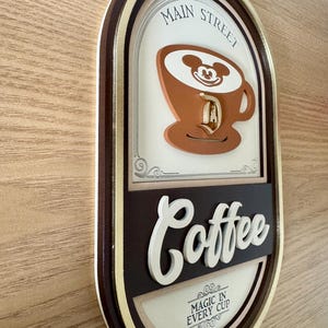 May include: A brown and gold oval sign with the text "Main Street Coffee" and a cartoon image of Mickey Mouse in a coffee cup. The sign also says "Magic in Every Cup".