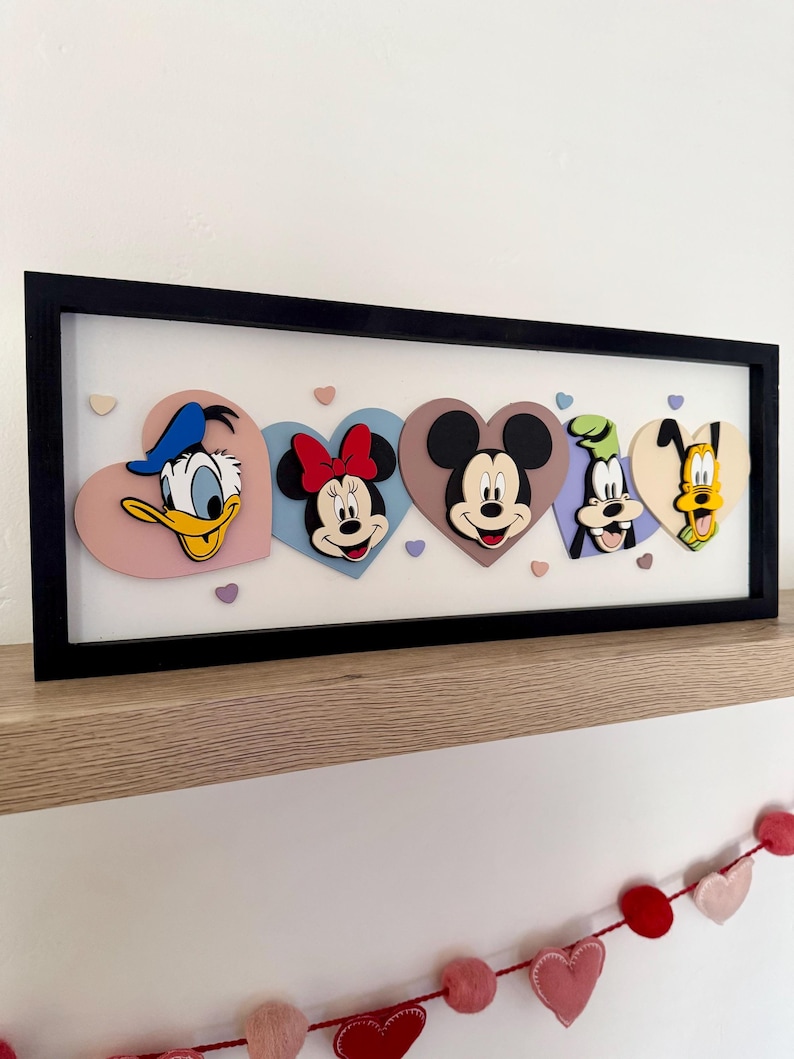 May include: A framed artwork featuring Disney characters Donald Duck, Minnie Mouse, Mickey Mouse, Goofy, and Pluto, each on a heart-shaped background. The characters are in a black frame, with a white background. The artwork is displayed on a wooden shelf.