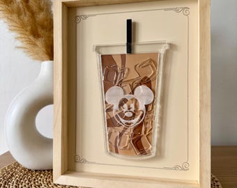 Könnte beinhalten: Eine gerahmte Illustration eines Mickey-Mouse-Kopfes in einem durchsichtigen Plastikbecher, gefüllt mit brauner Flüssigkeit und Eis. Der Becher hat einen schwarzen Strohhalm und ist vor einem cremefarbenen Hintergrund platziert.