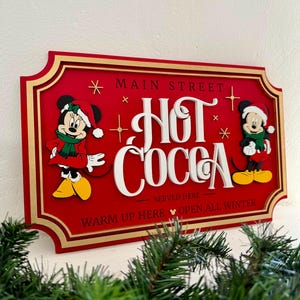 K&ouml;nnte beinhalten: Ein rotes Holzschild mit goldenen Verzierungen, auf dem Mickey und Minnie Mouse in Winterkleidung zu sehen sind. Das Schild lautet "Main Street Hot Cocoa Served Here Warm Up Here Open All Winter".