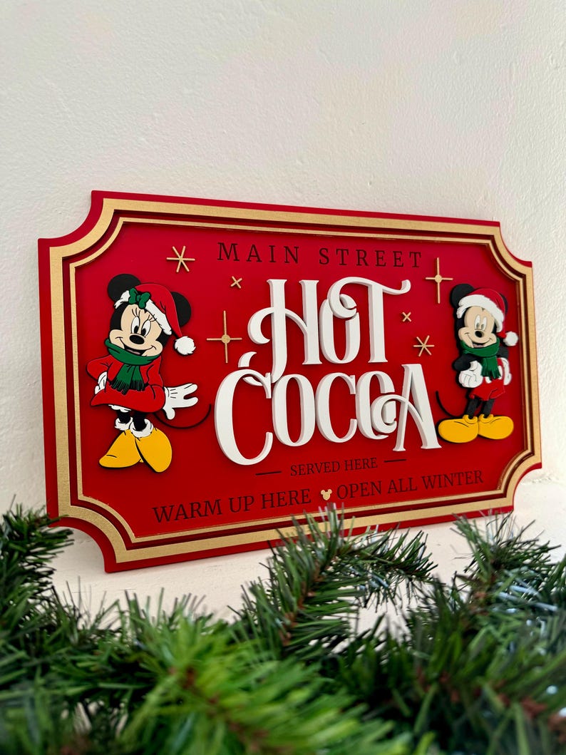 K&ouml;nnte beinhalten: Ein rotes Holzschild mit goldenen Verzierungen, auf dem Mickey und Minnie Mouse in Winterkleidung zu sehen sind. Das Schild lautet "Main Street Hot Cocoa Served Here Warm Up Here Open All Winter".