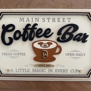 K&ouml;nnte beinhalten: Ein wei&szlig;es Schild mit schwarzer Beschriftung, auf dem "Main Street Coffee Bar" steht. Das Schild zeigt eine braune Kaffeetasse mit dem Gesicht von Mickey Mouse im Schaum und einem Disney-Schlosslogo an der Seite. Das Schild zeigt auch "Fresh Coffee", "Open Daily", "Since 1955" und "A Little Magic in Every Cup".
