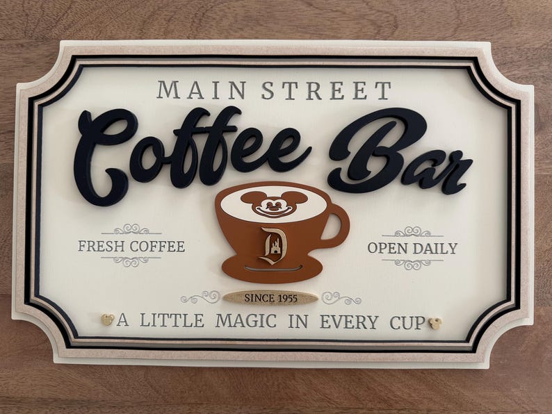 K&ouml;nnte beinhalten: Ein wei&szlig;es Schild mit schwarzer Beschriftung, auf dem "Main Street Coffee Bar" steht. Das Schild zeigt eine braune Kaffeetasse mit dem Gesicht von Mickey Mouse im Schaum und einem Disney-Schlosslogo an der Seite. Das Schild zeigt auch "Fresh Coffee", "Open Daily", "Since 1955" und "A Little Magic in Every Cup".