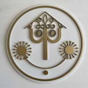May include: A gold-colored metal wall decor with a stylized design of a house with a whimsical roof and two dandelion-like shapes on either side. The design is surrounded by a gold-colored circle.