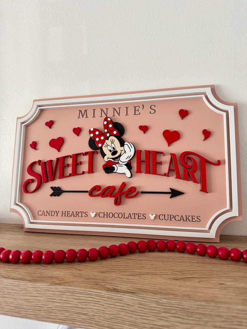May include: A pink and white wooden sign with a red and white Minnie Mouse illustration. The sign says "Minnie's Sweetheart cafe" with an arrow pointing to the right. The text below the arrow says "Candy Hearts Chocolates Cupcakes".