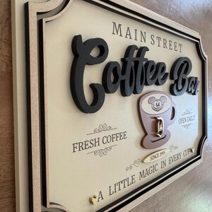 K&ouml;nnte beinhalten: Ein Holzschild mit einem 3D-Design, auf dem "Main Street Coffee Bar" steht. Das Schild ist mit einer braunen Kaffeetasse mit einem Mickey-Maus-Kopf verziert und dem Text "Fresh Coffee", "Open Daily", "Since 1955" und "A Little Magic in Every Cup".