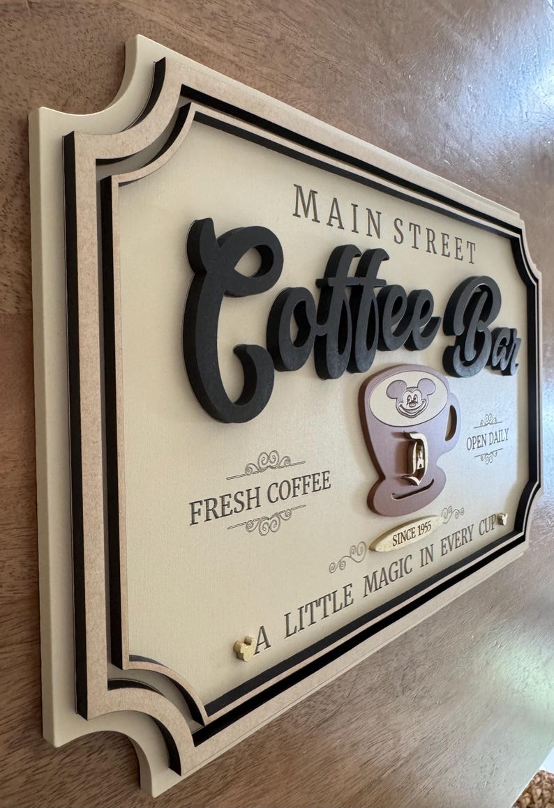 K&ouml;nnte beinhalten: Ein Holzschild mit einem 3D-Design, auf dem "Main Street Coffee Bar" steht. Das Schild ist mit einer braunen Kaffeetasse mit einem Mickey-Maus-Kopf verziert und dem Text "Fresh Coffee", "Open Daily", "Since 1955" und "A Little Magic in Every Cup".