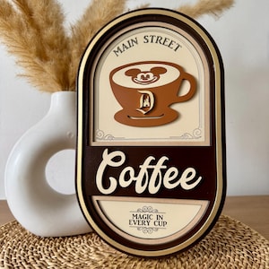 May include: A brown wooden sign with a gold frame and the text "Main Street Coffee" in white lettering. The sign features a cartoon image of Mickey Mouse's head in a cup of coffee with a Disney castle logo. The text "Magic in Every Cup" is also on the sign.
