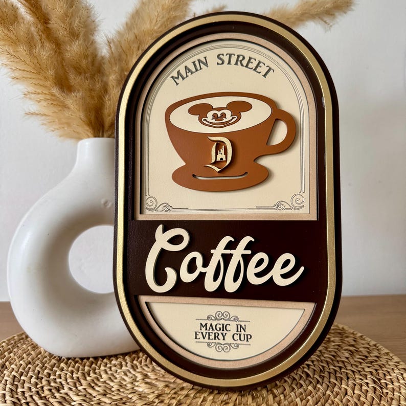 May include: A brown wooden sign with a gold frame and the text "Main Street Coffee" in white lettering. The sign features a cartoon image of Mickey Mouse's head in a cup of coffee with a Disney castle logo. The text "Magic in Every Cup" is also on the sign.