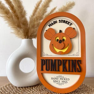 May include: Orange wooden sign with a Mickey Mouse pumpkin face and the text "Main Street Pumpkins Hand Picked Since 1955".